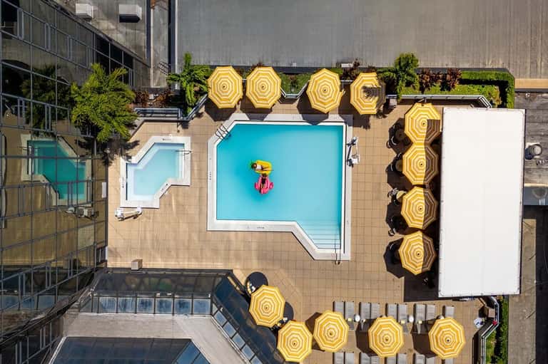Aerial view of rooftop pool with yellow umbrellas, turquoise water, and surrounding urban high-rise buildings