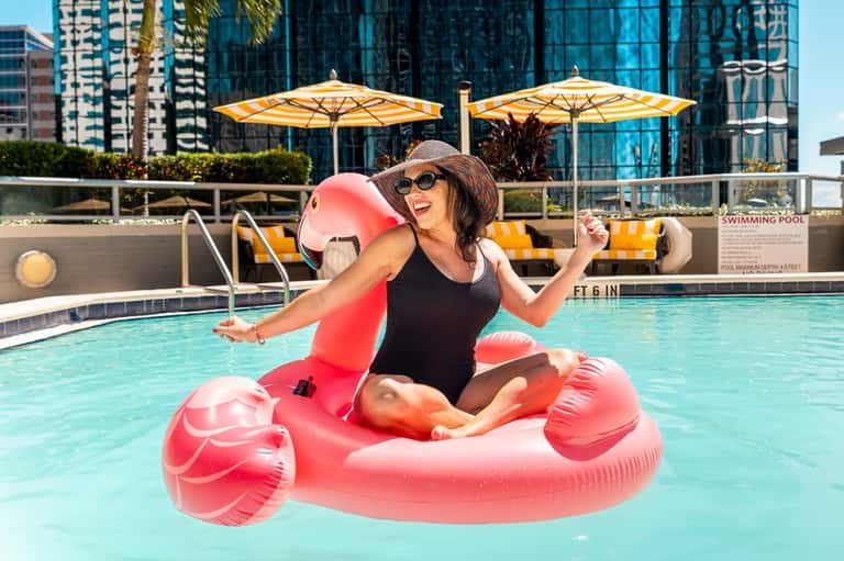 Woman in pool on pink flamingo float wearing hat and sunglasses with yellow umbrellas in background