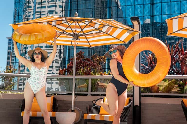 Two women by rooftop pool holding yellow floats under yellow and white striped umbrella with city skyline behind