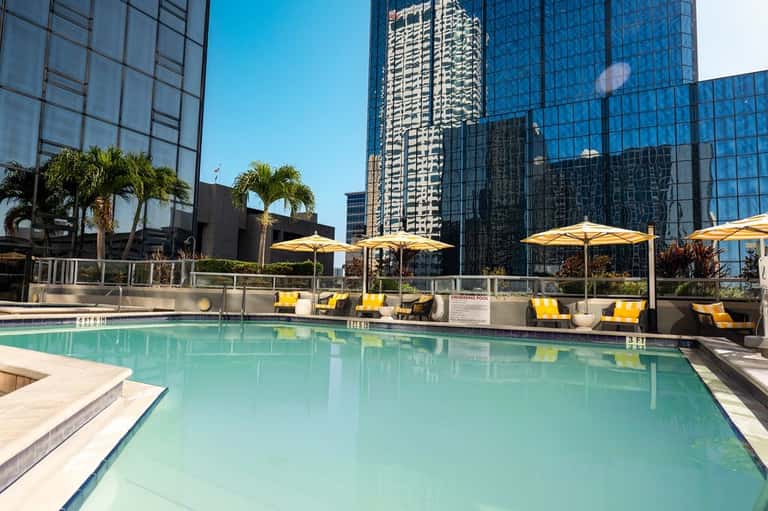 Rectangular pool with clear turquoise water, yellow umbrellas, lounge chairs, and modern city buildings surrounding