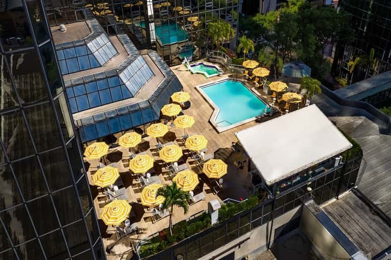 Aerial view of multi-level rooftop pool complex with yellow umbrellas, loungers, and surrounding glass buildings