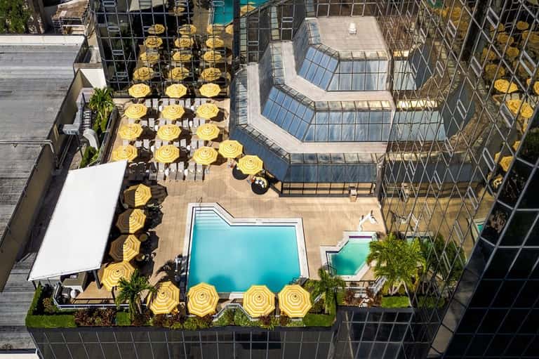 Top-down view of urban rooftop pool area with turquoise water, yellow umbrellas, lounge chairs, and tall buildings