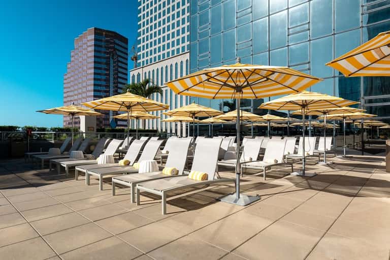 Rooftop deck with yellow and white striped umbrellas, lounge chairs, and modern high-rise buildings under clear blue sky