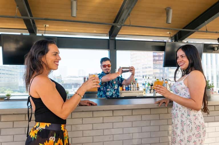Three people at an indoor bar with city skyline views, bartender pouring drinks and guests toasting with cocktails