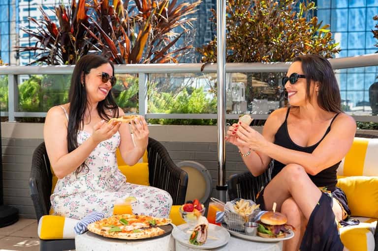 Two women dining outdoors on a rooftop terrace with city views, food and drinks on table, decorative plants in background