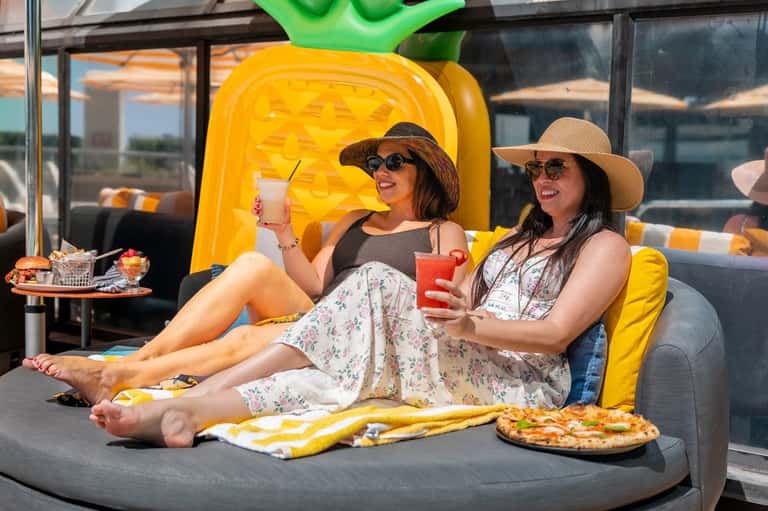 Two women relaxing on a rooftop lounge with yellow cushioned seating, pizza and drinks, wearing hats and sunglasses