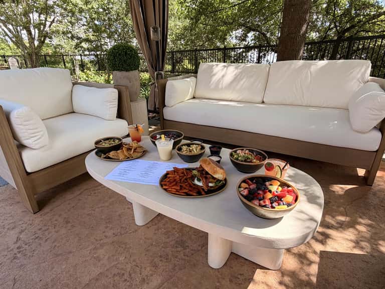 Upscale outdoor dining setup with cream sofa, armchair, food spread on white table, and wooded backdrop