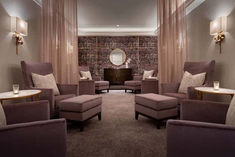 Elegant spa lounge with symmetrical purple armchairs, ottomans, brass wall sconces, and dark accent wall