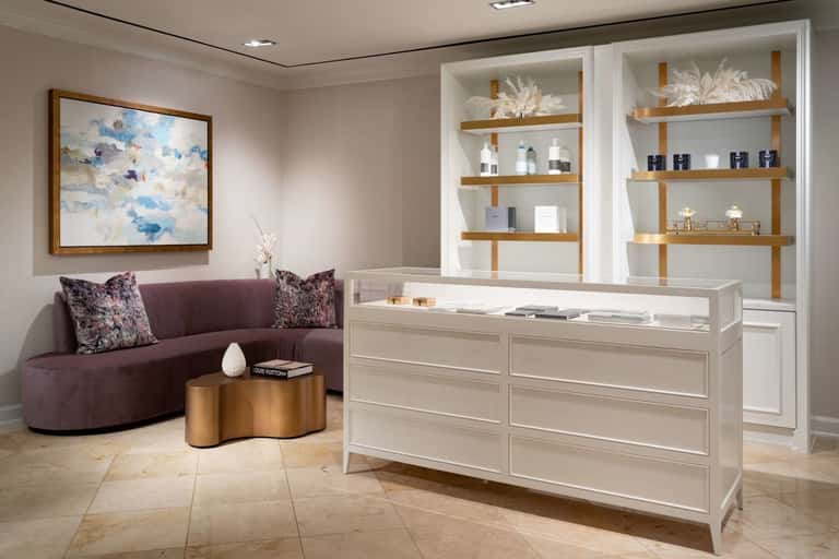 Modern spa reception area with curved plum sofa, gold side table, and white display shelving with products