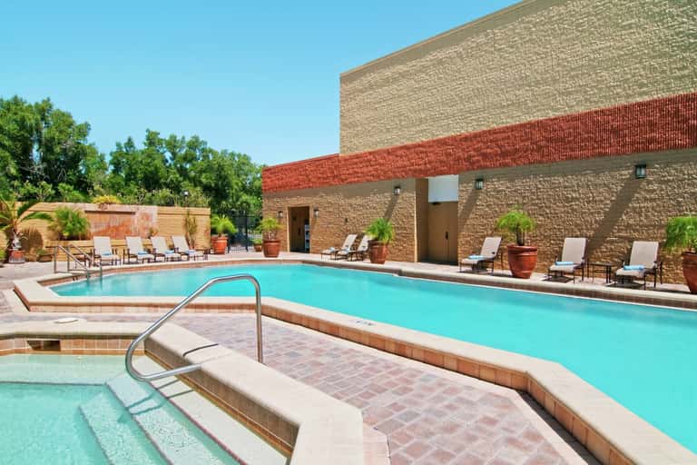 Outdoor swimming pool with lounge chairs, brick building, palm trees, and clear blue sky