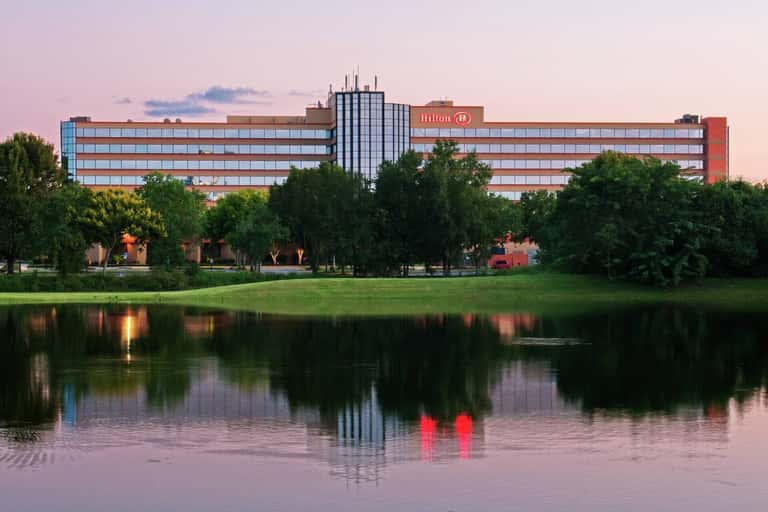 Hilton hotel exterior at dusk, multi-story building reflected in calm water, surrounded by landscaped grounds and trees