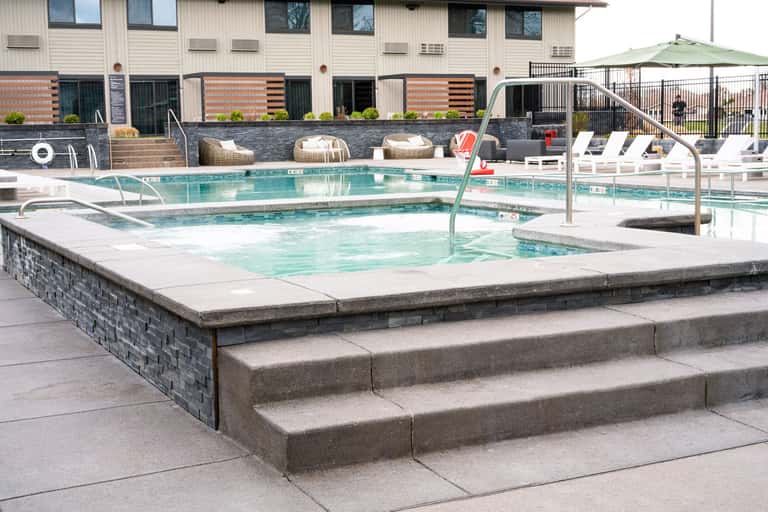 Outdoor hot tub and swimming pool area with lounge chairs, umbrella, and modern hotel building in background