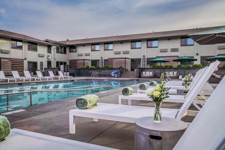 Resort pool area with lap lanes, white lounge chairs, rolled towels, flower arrangements, and modern building with green umbrellas
