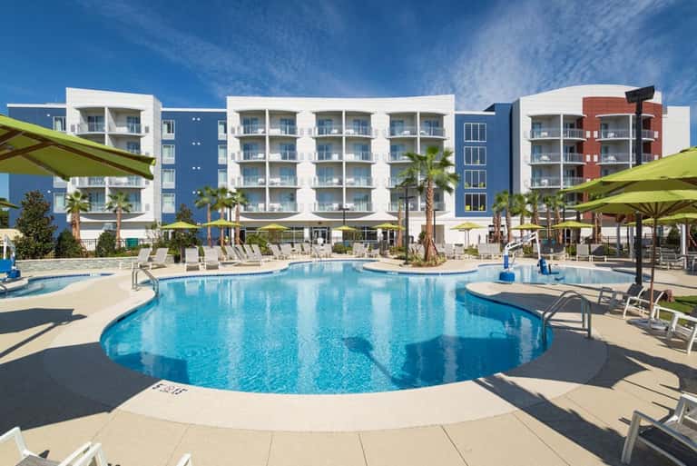 Resort pool with curved edges surrounded by lounge chairs, palm trees, and multi-story apartment buildings