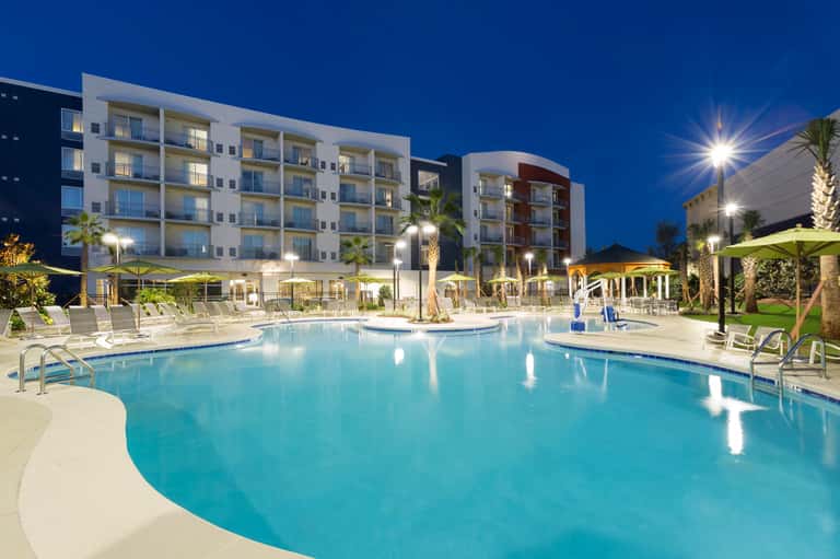 Illuminated resort pool at dusk with loungers, yellow umbrellas, lit buildings, and palm trees