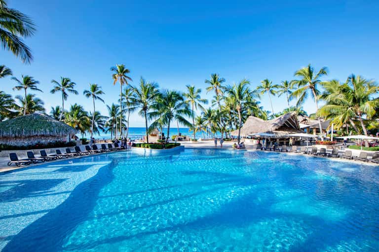 Tropical resort pool with crystal-clear turquoise water, palm trees, and thatched-roof structures