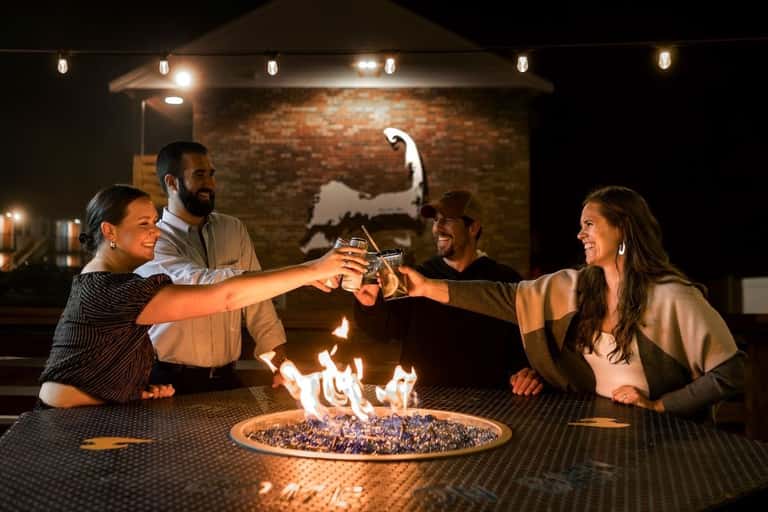 Four friends toasting with drinks around a fire pit on a patio at night