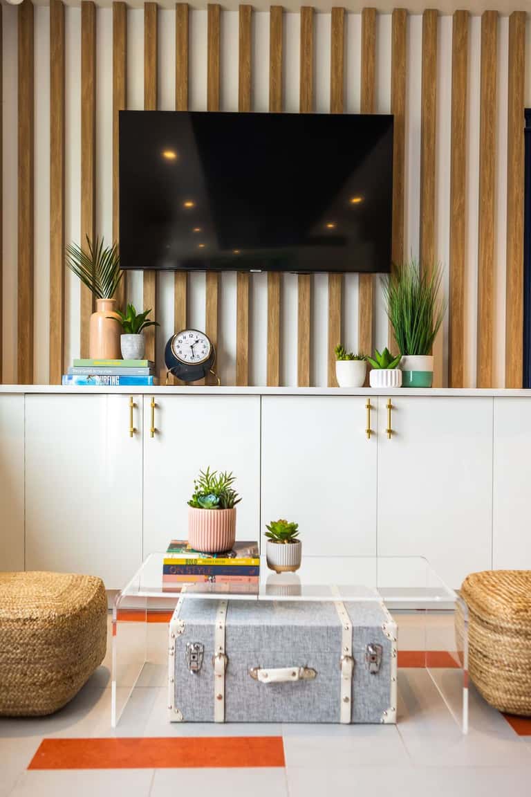 Modern living room with wood slat wall, mounted TV, white storage cabinets, and woven ottomans