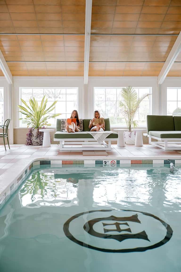 Indoor pool facility with wooden ceiling, large windows, green seating, potted plants, and natural light