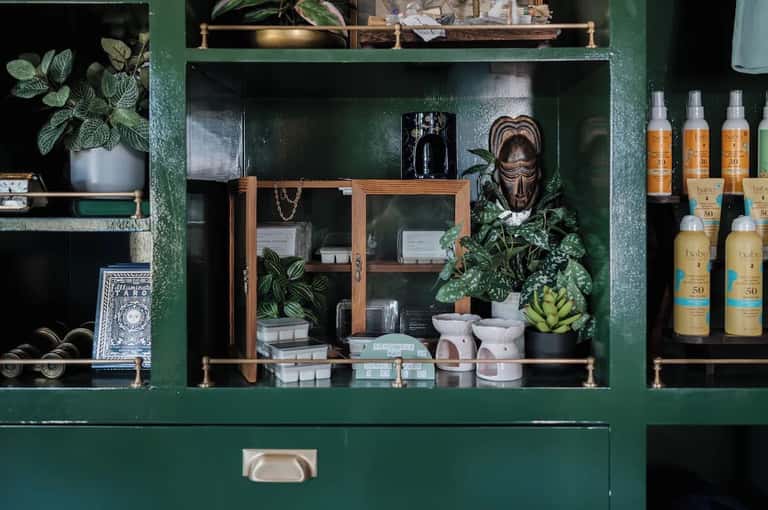 Green retail display cabinet with potted plants, decorative items, sunscreen bottles, and wellness products on shelves
