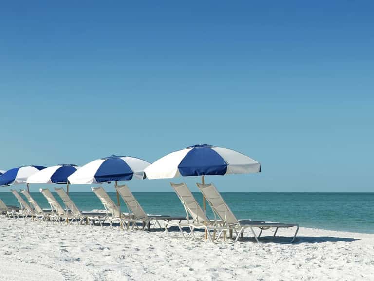 Row of blue and white beach umbrellas with lounge chairs on white sand facing calm turquoise water