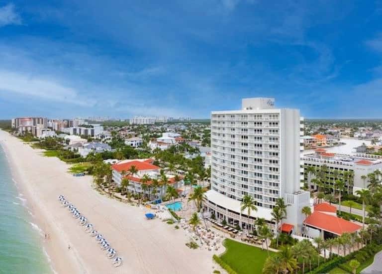 Aerial view of beachfront resort with multi-story white building, beach umbrellas, and palm trees along shore