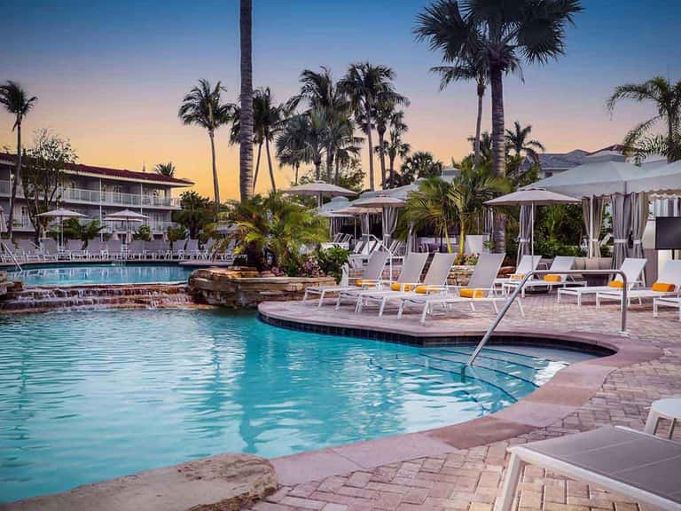 Resort pool area at dusk with lounge chairs, cabanas, and palm trees under twilight sky