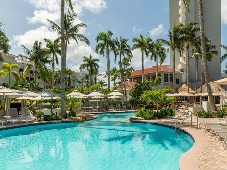 Large resort pool with cabanas, white umbrellas, palm trees, and multi-story buildings surrounding
