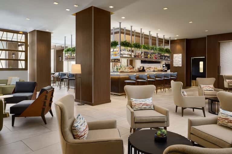Modern hotel lounge with brown wood columns, cream armchairs, colorful pillows, and bar with bottles in background