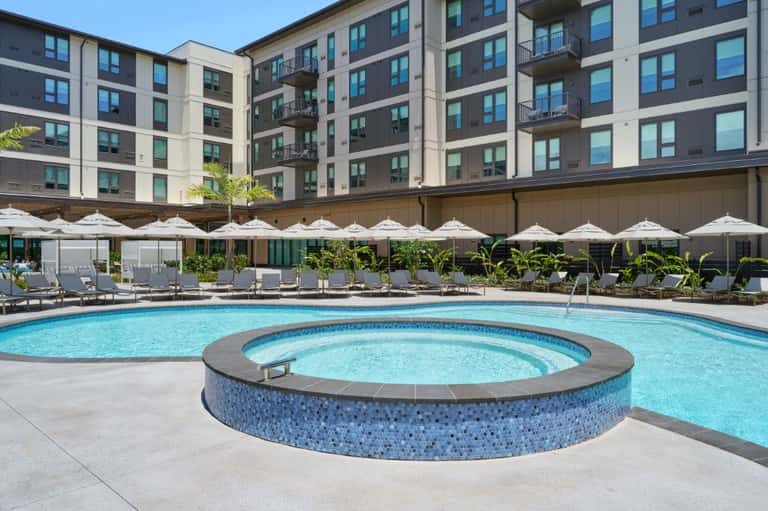 Hotel courtyard with main pool, hot tub, white umbrellas, lounge chairs, and multi-story residential building with balconies