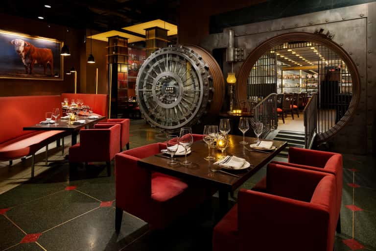 Upscale dining room with red seating, dark tables, industrial vault doors, and warm ambient lighting