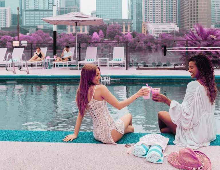 Two women in summer dresses toasting drinks by a pool with purple flowering trees and city skyline backdrop