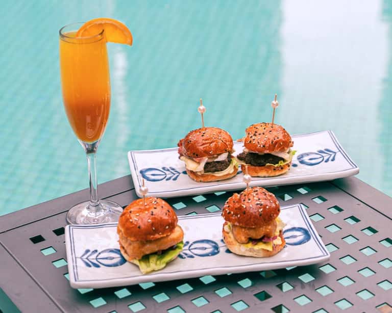 Four mini sliders with orange cocktail garnished with fruit on decorative white plates