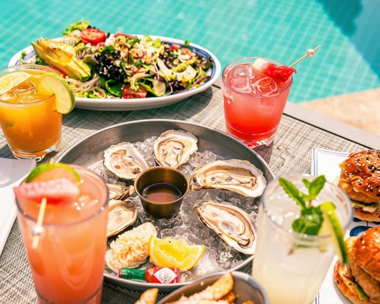 Poolside dining spread with oysters, colorful noodle salad, and tropical cocktails