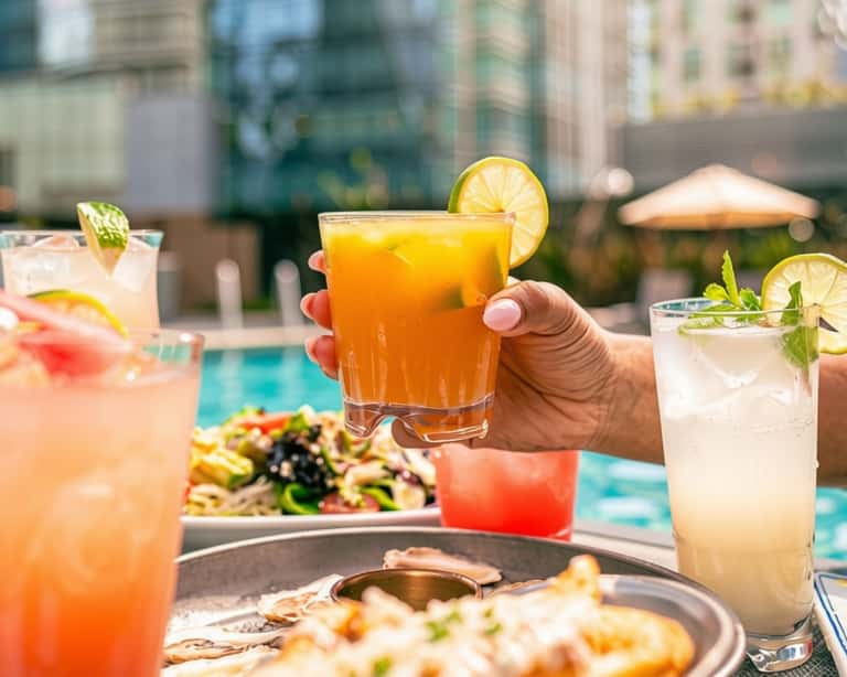 Person holding a tropical cocktail with multiple colorful drinks and appetizers visible