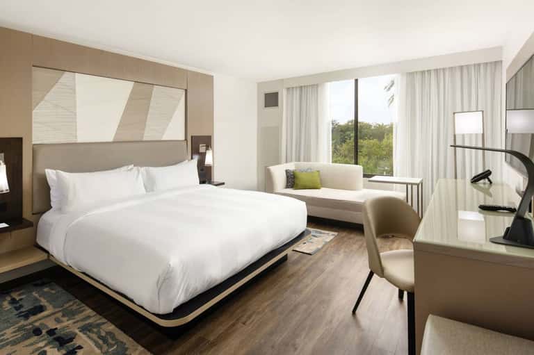 Upscale hotel bedroom featuring king bed, sofa seating area, desk, and large windows overlooking trees