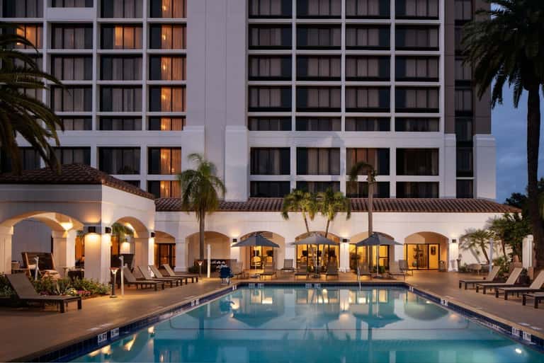 Resort swimming pool at dusk with lounge chairs, arched colonnade, and illuminated hotel towers