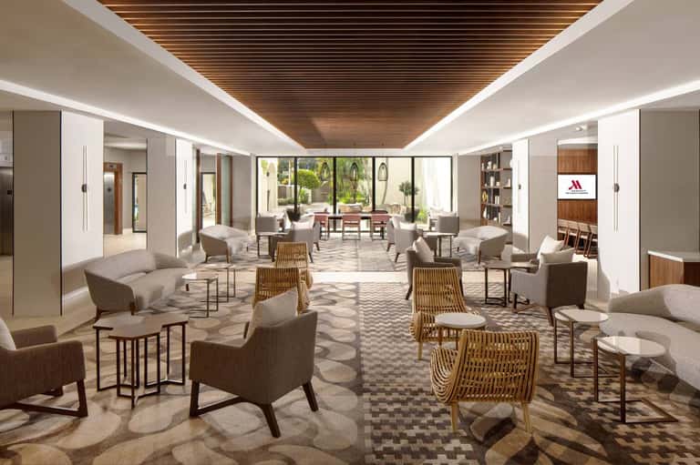 Modern hotel lounge with beige seating, wooden chairs, patterned flooring, and large windows overlooking courtyard