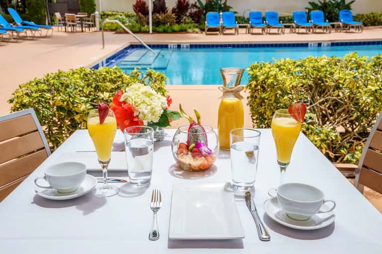 Poolside breakfast table with fresh juice, fruit, pastries, flowers, and ocean view in background