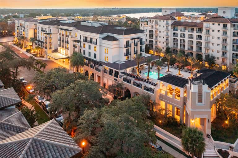 Aerial sunset view of luxury resort complex with illuminated buildings, pool, palm trees, and waterfront