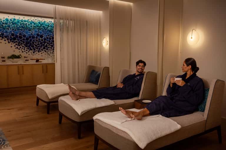 Relaxation lounge with two guests in spa robes sitting on beige chaise lounges with modern lighting