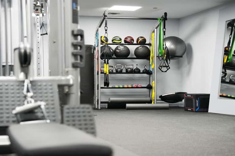 Modern gym facility with organized equipment shelves, medicine balls, kettlebells, suspension trainers, and cardio machines