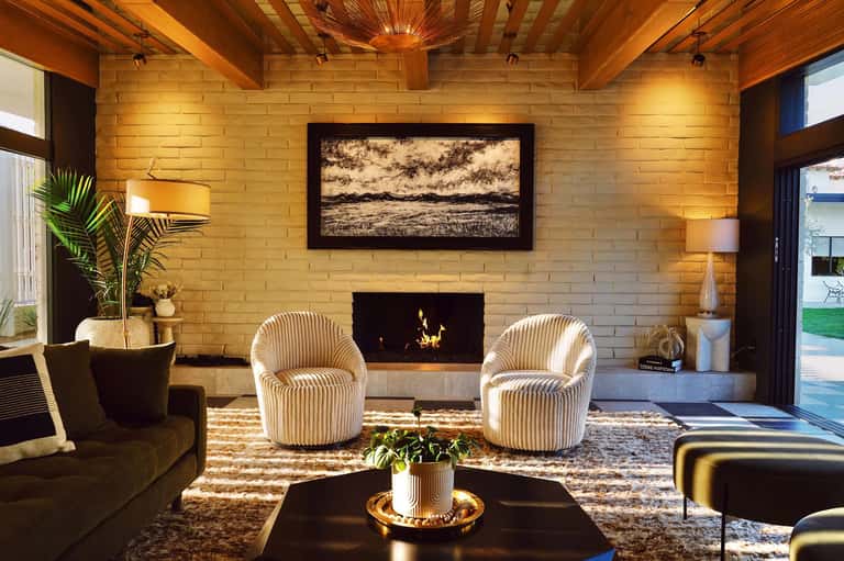 Luxury living room with brick fireplace, cream barrel chairs, wooden beams, and modern artwork