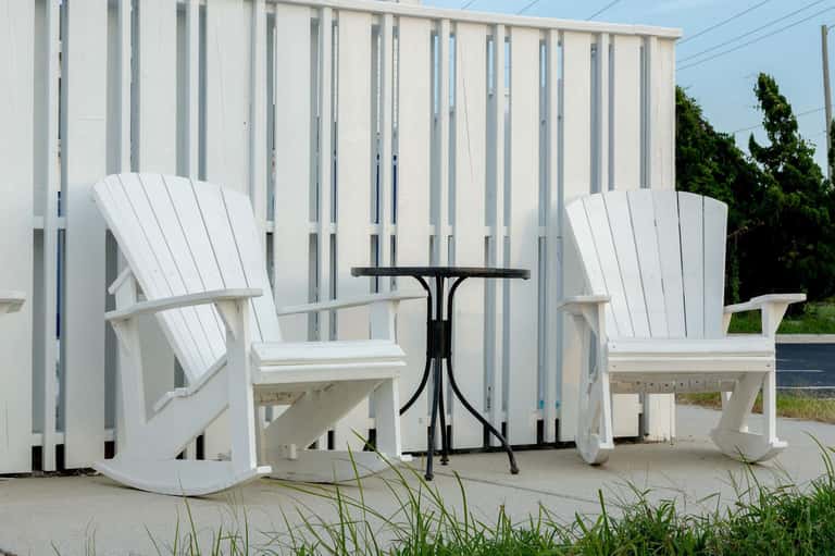 Two white Adirondack chairs on sandy patio with white fence and black side table