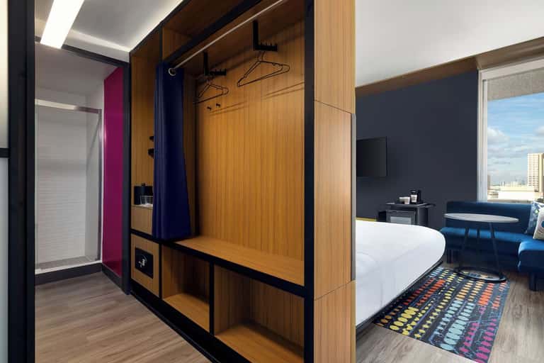 Modern hotel room with wooden wardrobe, white bed, colorful geometric rug, and city view through large windows
