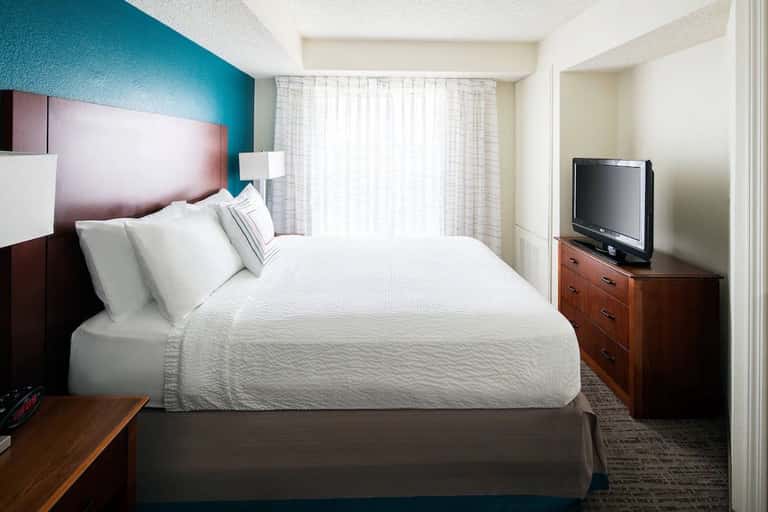 Modern hotel room featuring burgundy headboard, white quilted bedding, flat-screen TV, and natural light from windows