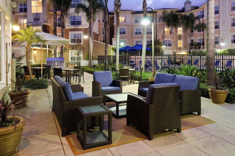 Outdoor patio seating area at night with rattan furniture, blue cushions, fire pit, and palm trees