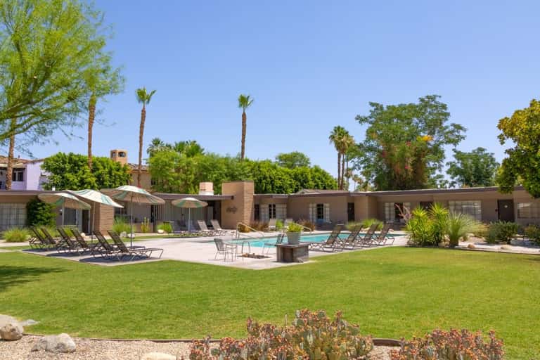 Resort courtyard with swimming pool, lounge chairs, umbrellas, and brick buildings surrounded by palm trees