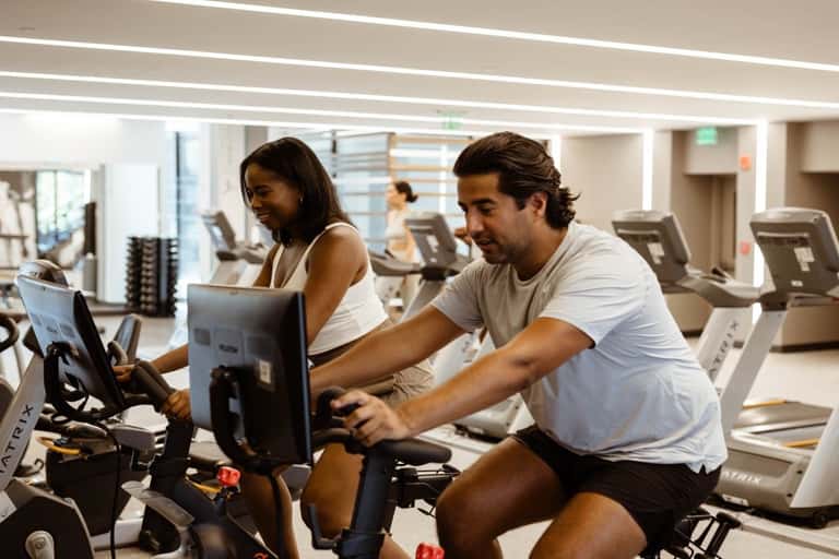 Two people exercising on stationary bikes in a bright, modern gym with multiple cardio machines