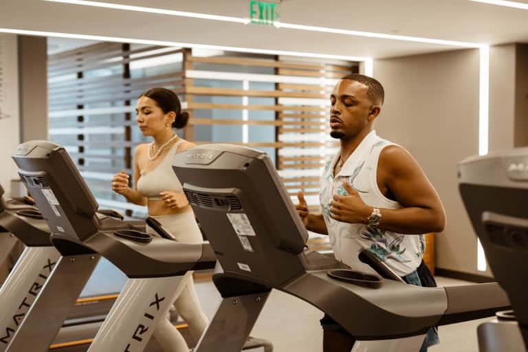 Man and woman running on treadmills in a well-lit fitness facility with wooden slat accents